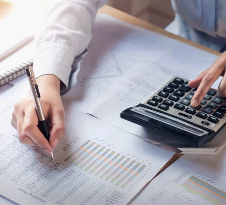 A person uses a calculator and writes on financial documents with charts and graphs on a desk, suggesting work related to accounting or data analysis.