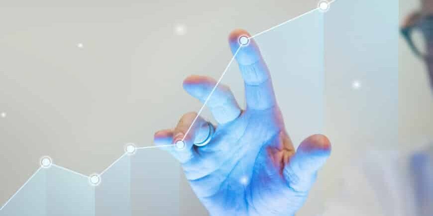A person’s hand touches a glowing upward-trending line graph, symbolizing growth or success. The background is blurred, and only part of the person’s face and glasses are visible.